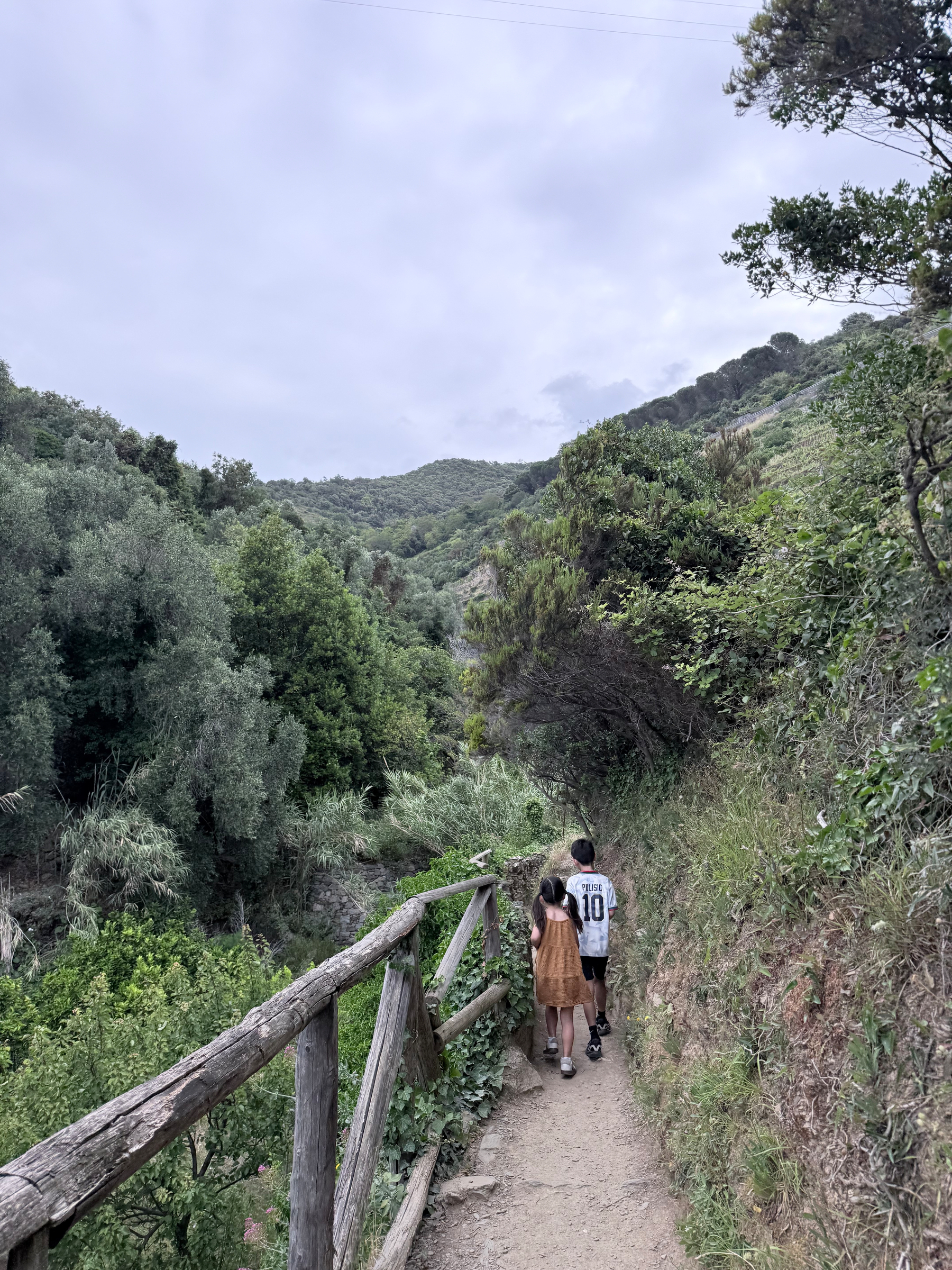 Two people walking along a narrow path surrounded by lush greenery and hills.