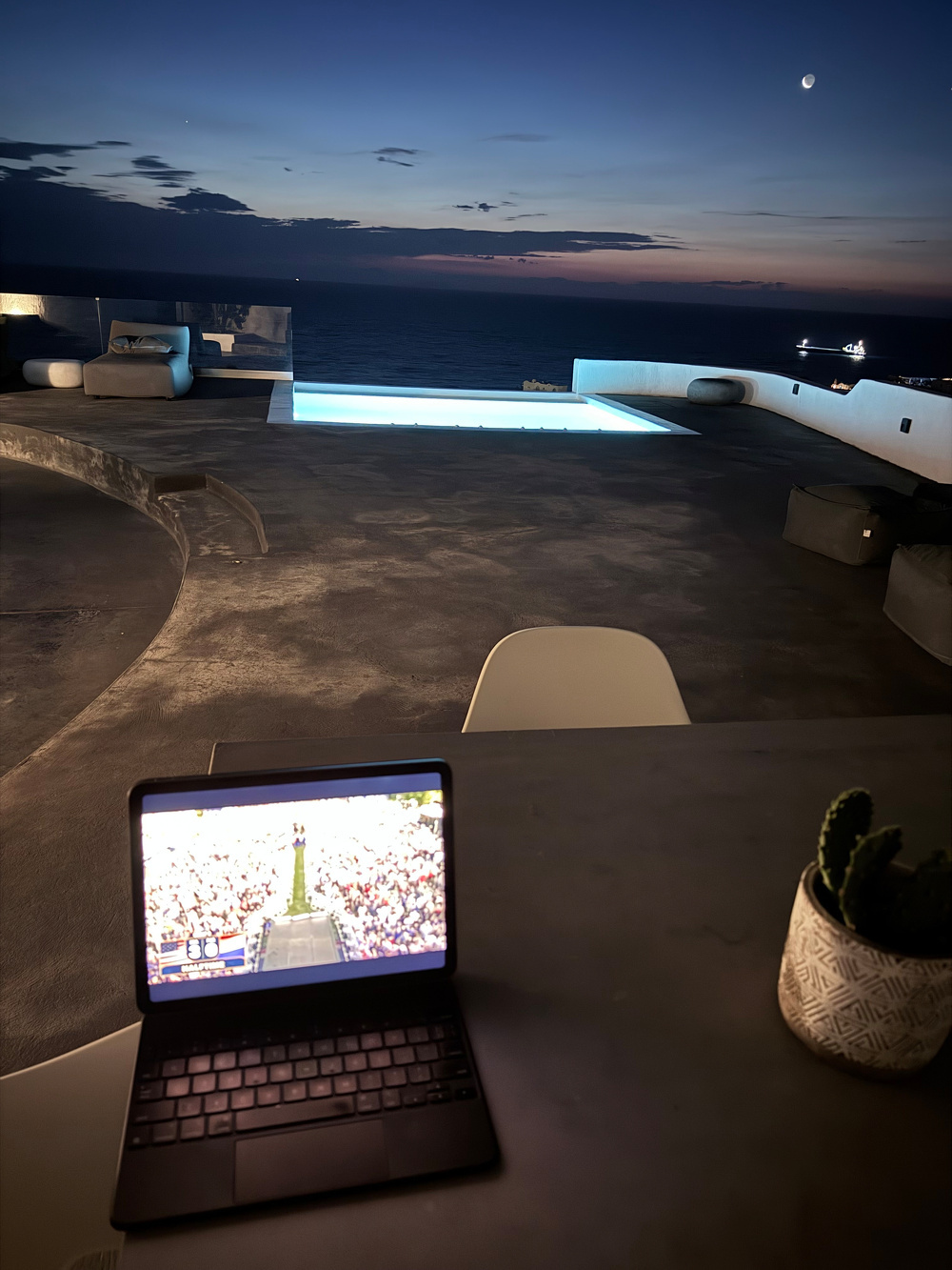 A tablet showing a concert sits on a table overlooking a scenic nighttime poolside view with a crescent moon in the sky.