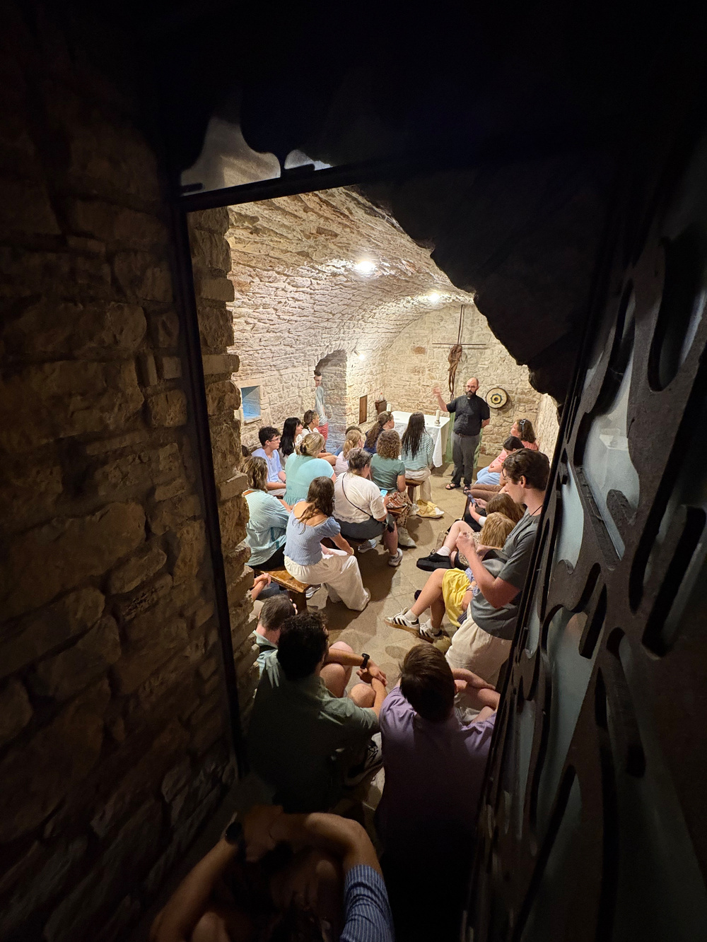 A group of people is seated in a rustic, stone-walled room, attentively listening to a speaker.