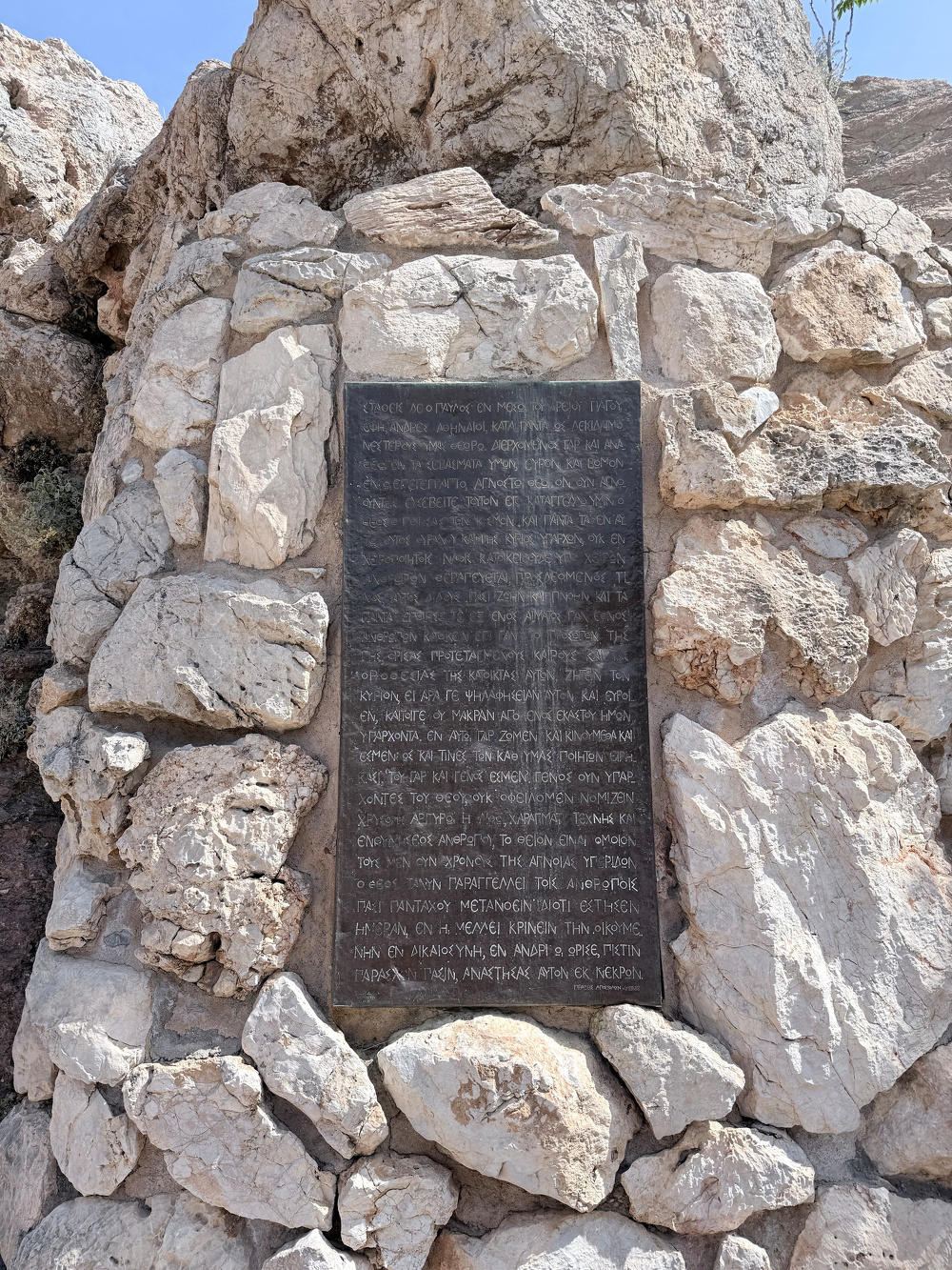 A stone slab with a plaque featuring ancient Greek text is mounted on a rock wall.