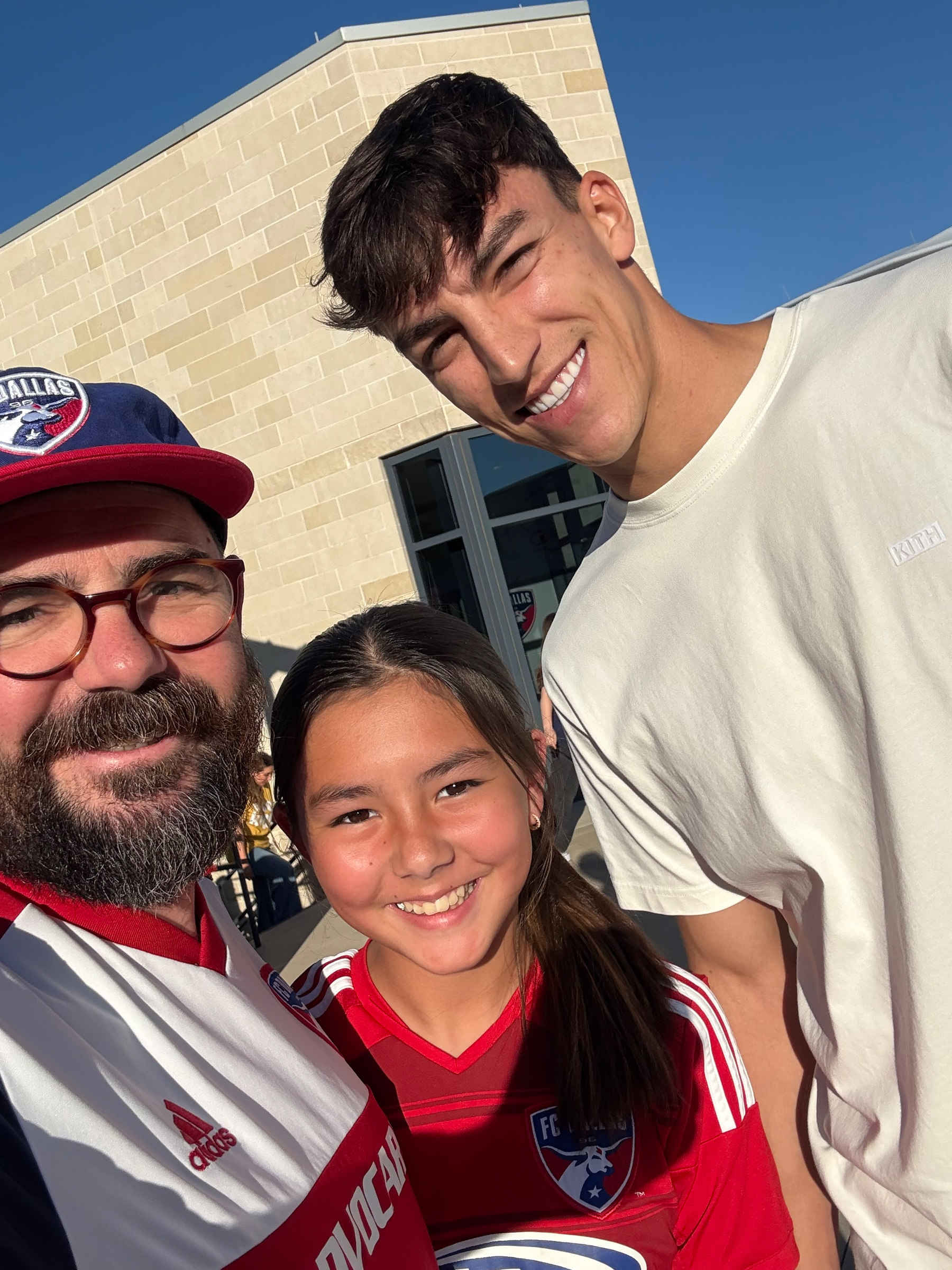 Three people are posing and smiling outdoors, with two of them wearing FC Dallas soccer jerseys.
