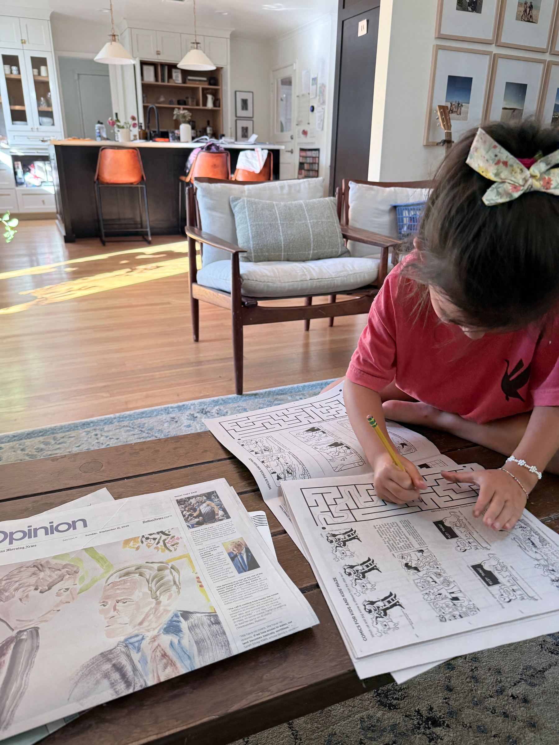 A young girl is focused on solving a maze puzzle in a newspaper while sitting at a wooden coffee table in a cozy living room.