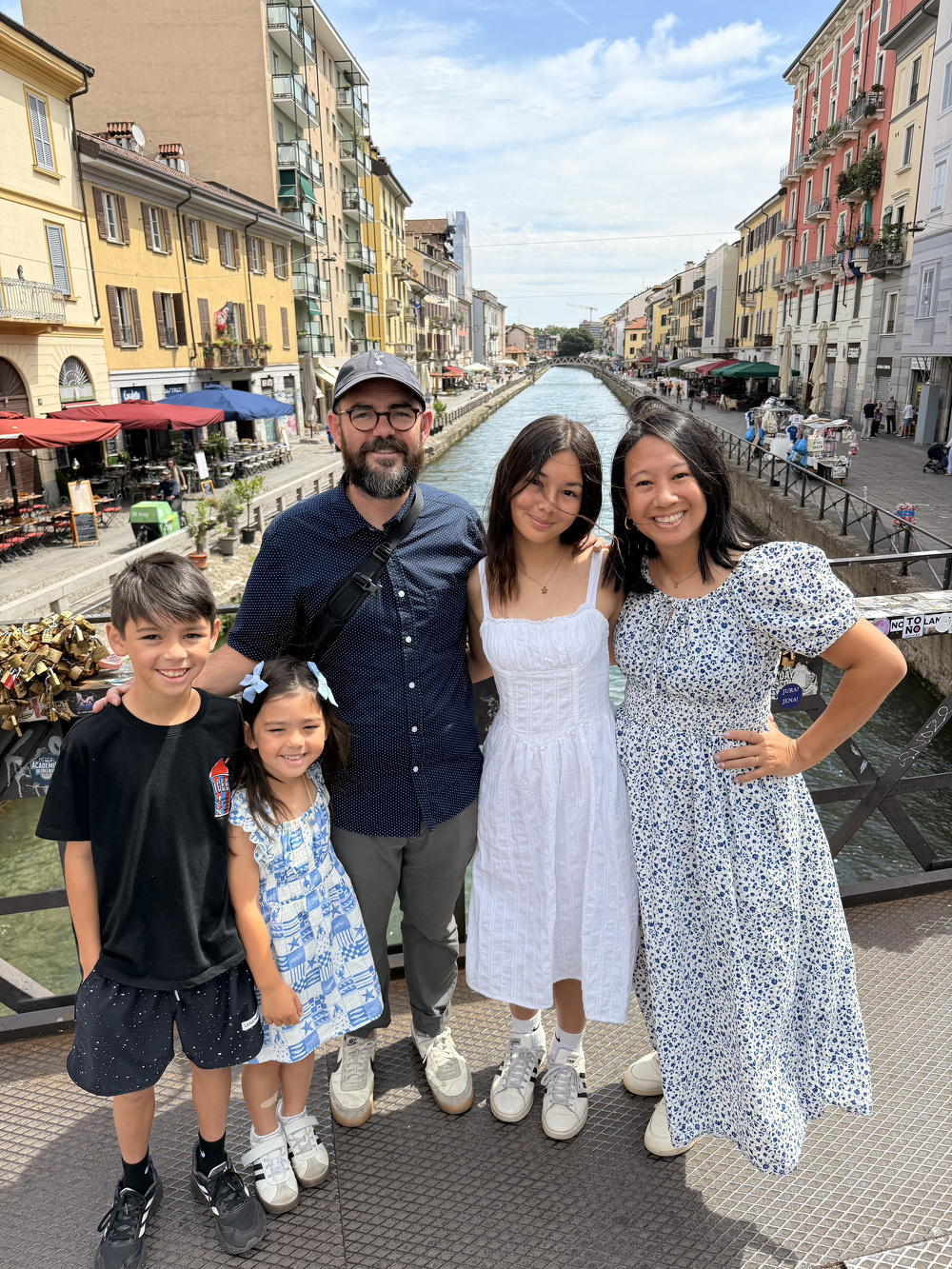A family of five poses happily on a bridge over a canal in a sunny, picturesque urban setting with colorful buildings and outdoor cafes.