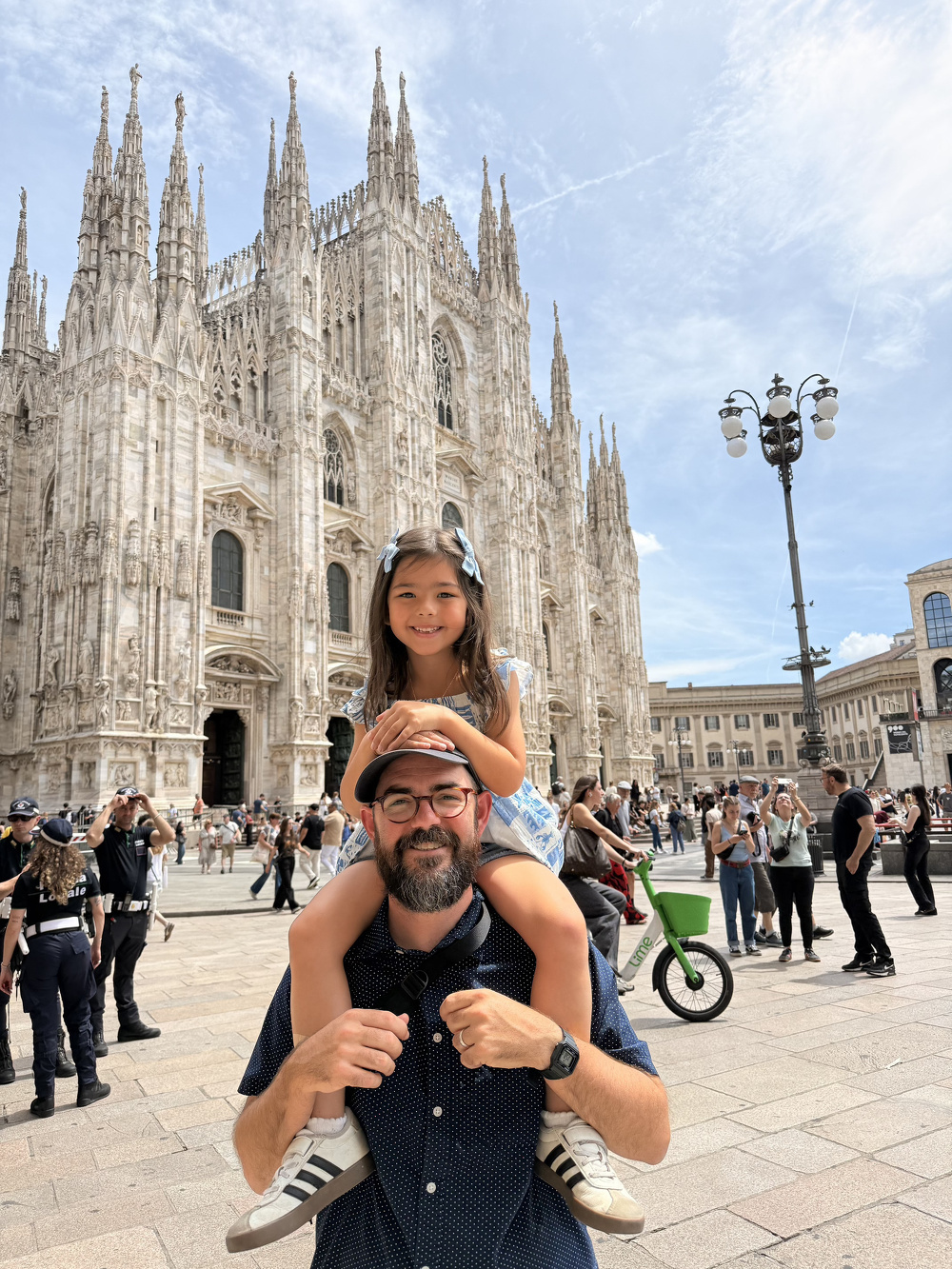 A man wearing glasses and a hat carries a smiling young girl on his shoulders in front of the ornate facade of the Milan Cathedral while a crowd gathers around them.