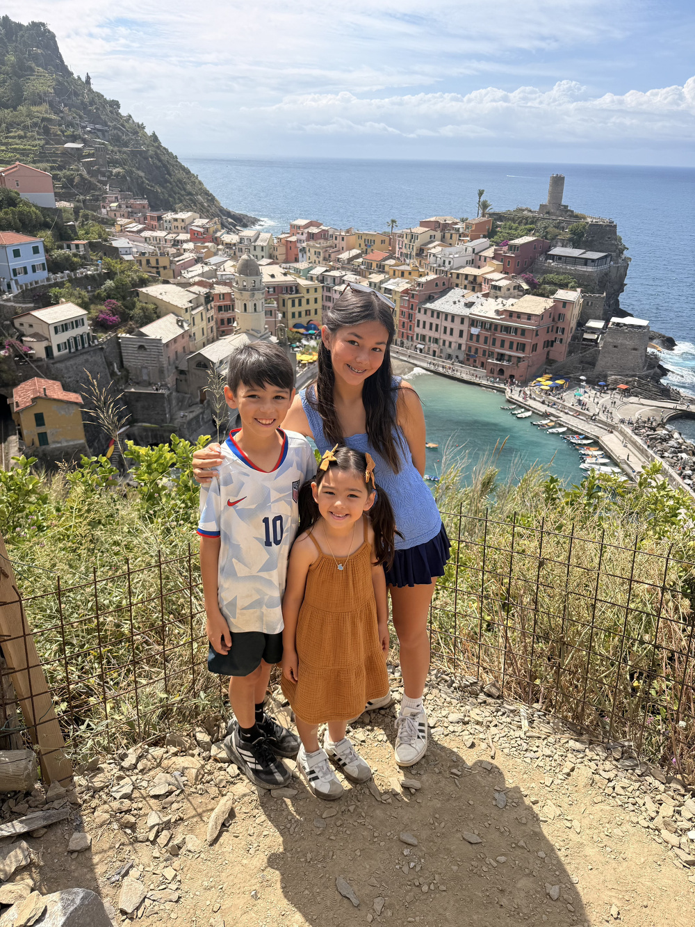 Three children pose happily on a hillside with a picturesque coastal village and ocean in the background.