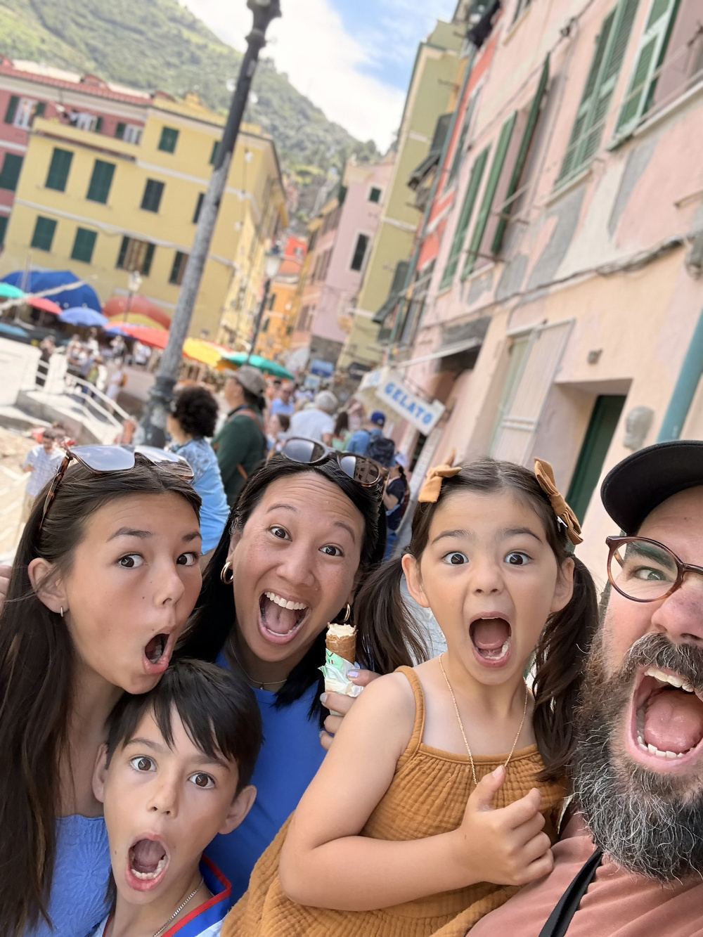 A family of five is joyfully posing with open mouths in a colorful, bustling outdoor market or street setting with buildings and umbrellas in the background.