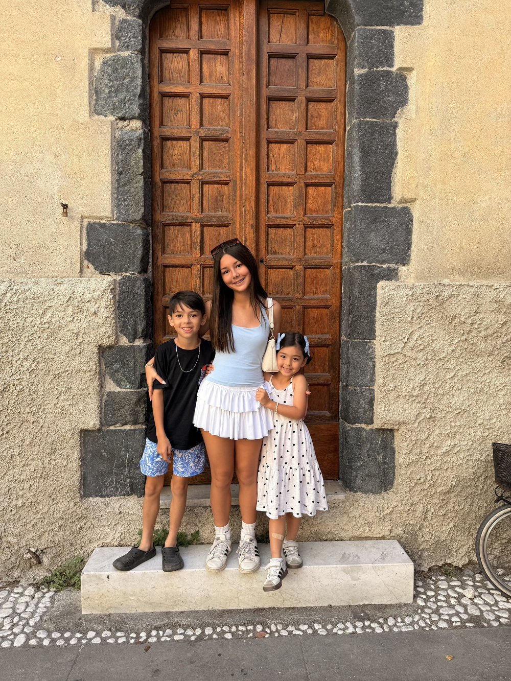 Three children are standing and smiling together in front of a large wooden door with stone trim.