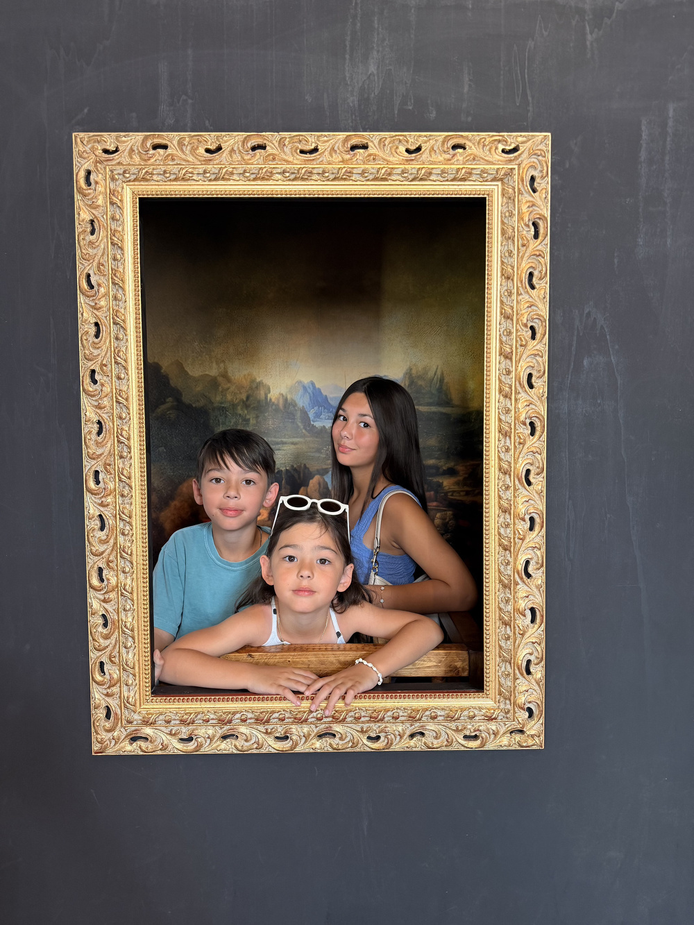 Three children are posing inside a large ornate picture frame set against a dark wall, creating the illusion that they are part of the framed artwork.