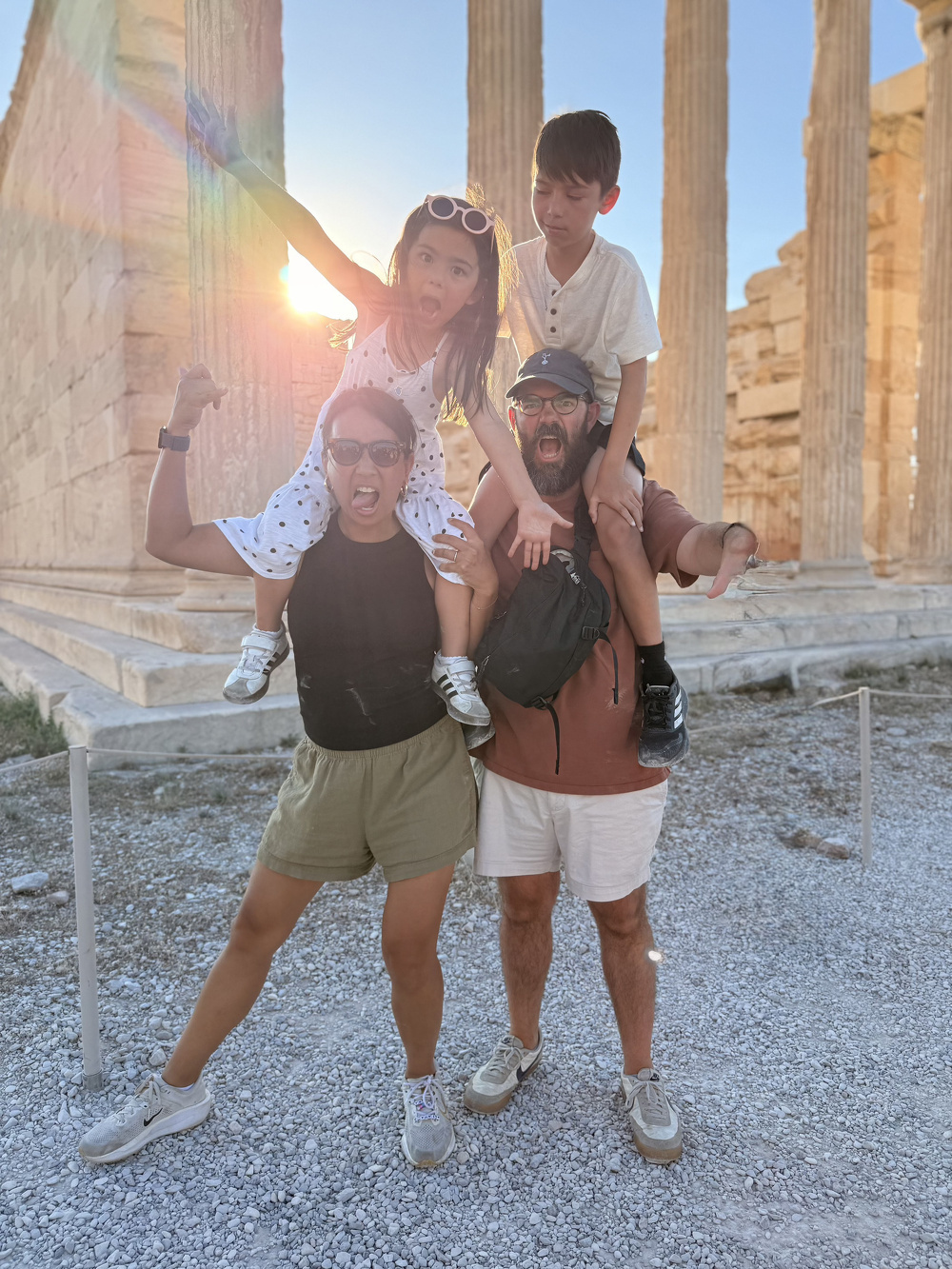 A family of four is posing playfully with two children sitting on the shoulders of the adults in front of ancient stone columns during sunset.