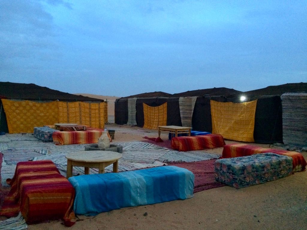 A cozy outdoor seating area is arranged with colorful cushions, rugs, and low tables under a dusky sky.