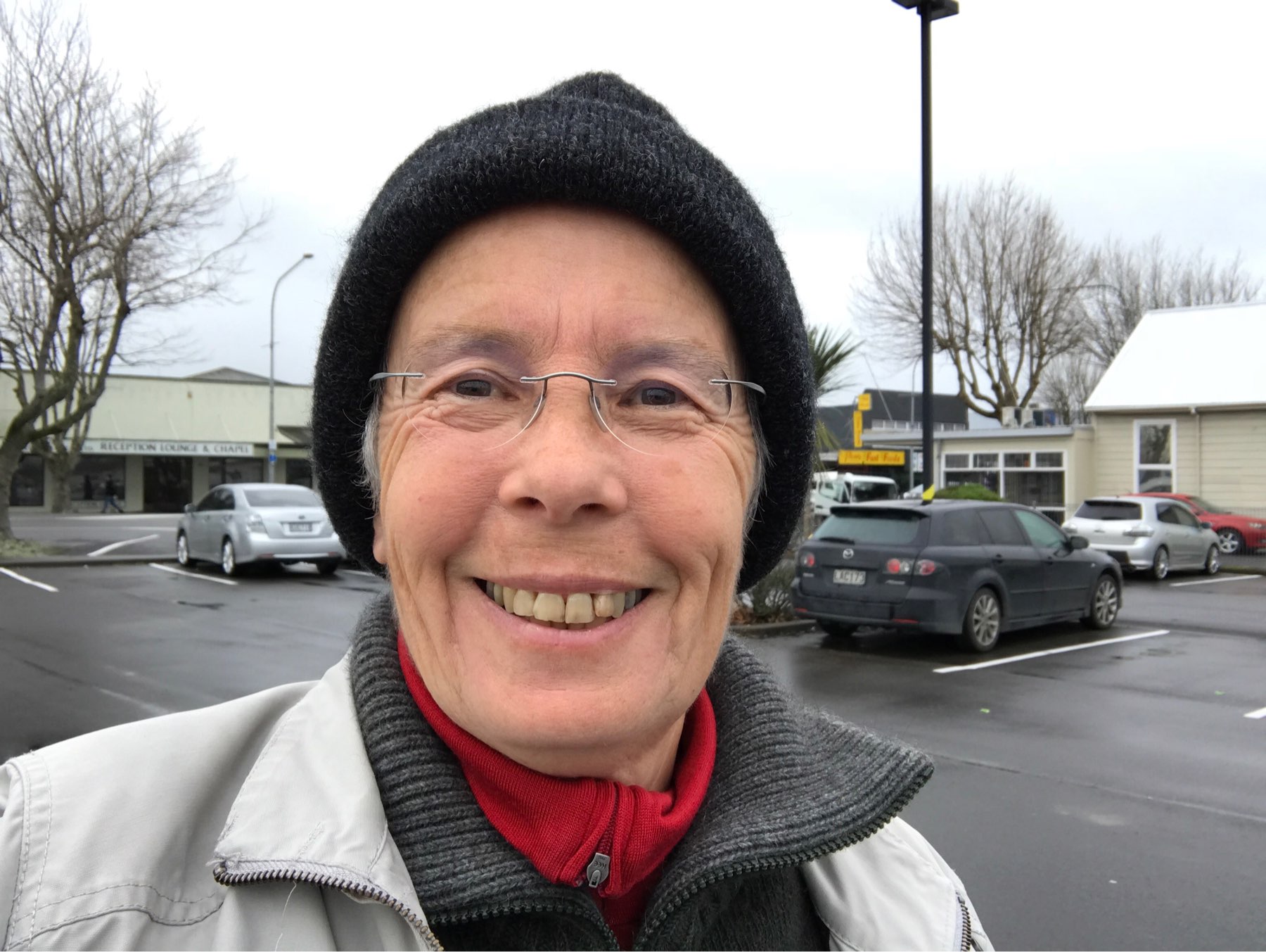 A person wearing glasses, a black beanie, and a light-colored jacket smiles at the camera in a parking lot on a cloudy day.