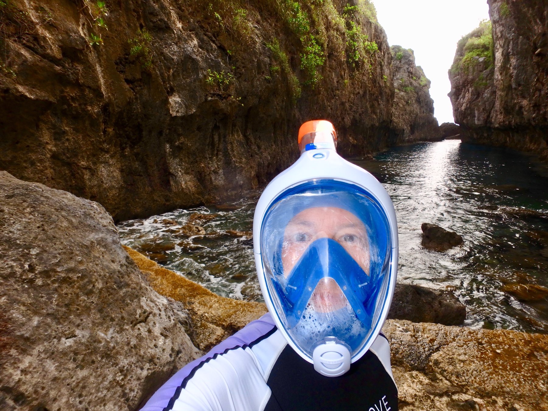 A person wearing a full-face snorkel mask stands on rocky terrain next to a narrow water passage between cliffs.