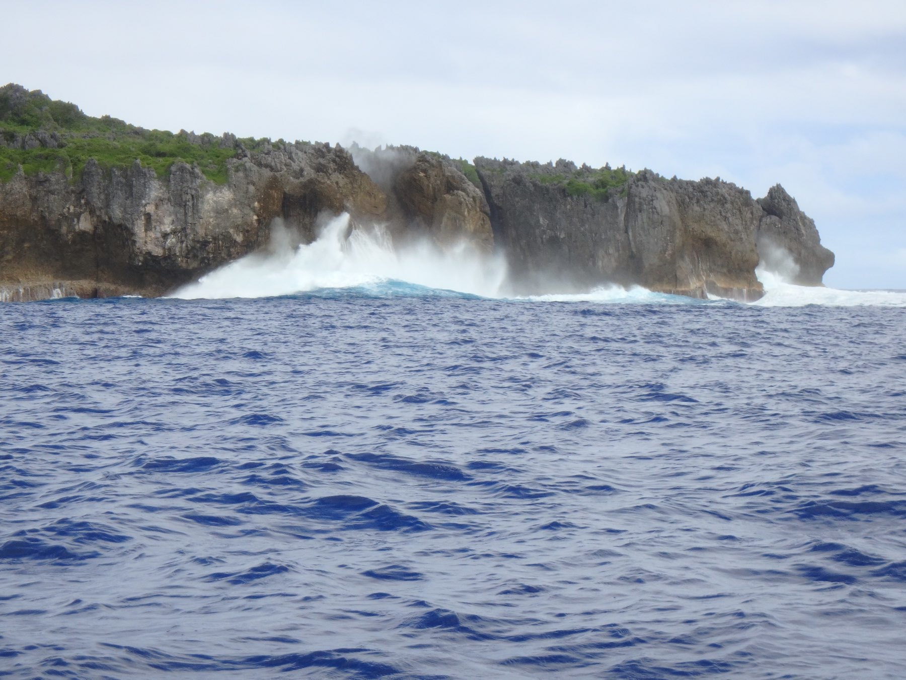 Waves are crashing against a rocky coastline under a cloudy sky.