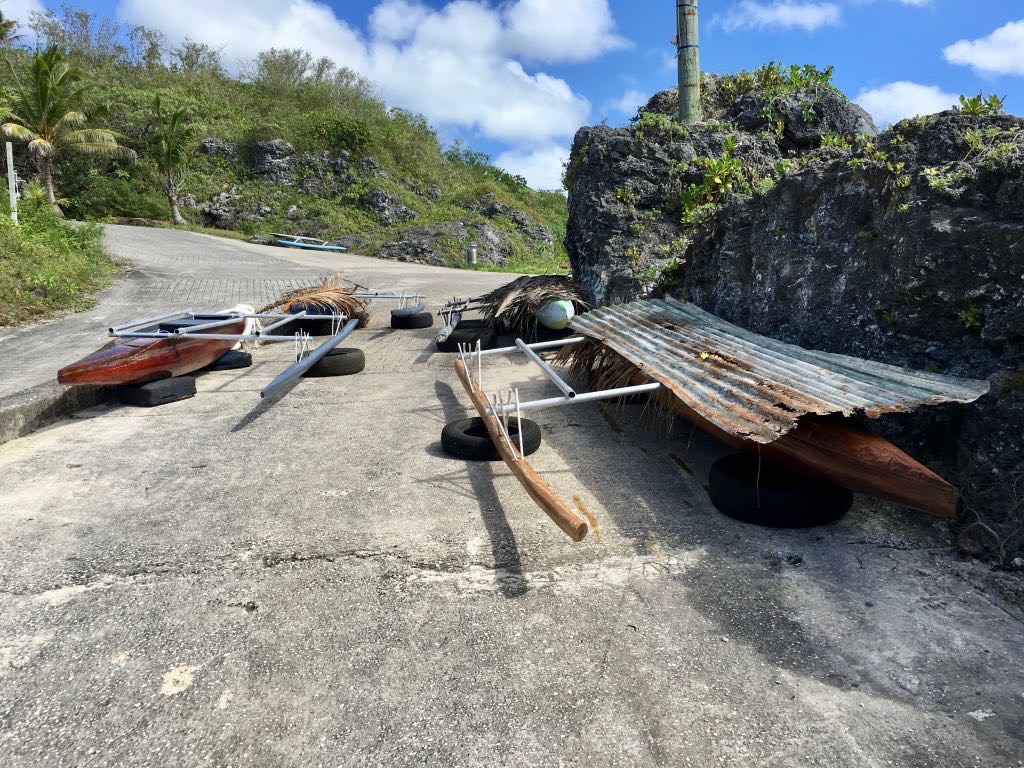 Two traditional outrigger canoes are resting on a concrete path near rocky terrain under a blue sky with scattered clouds.