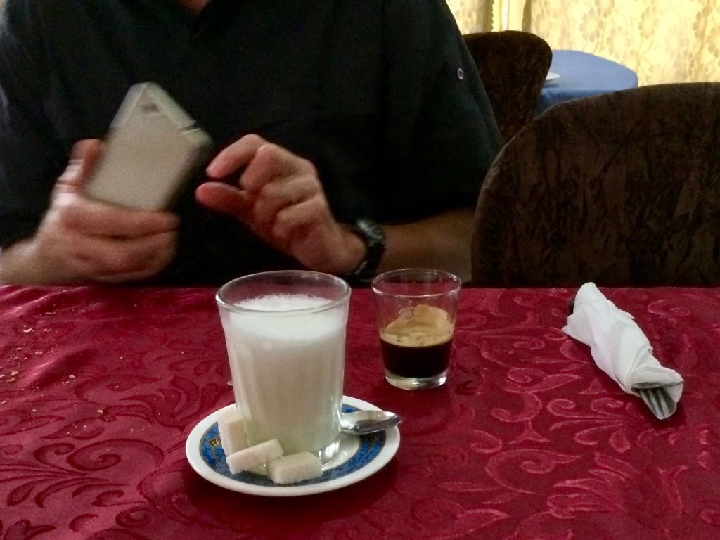A person is sitting at a table with a glass of milk, a small cup of espresso, and sugar cubes on a plate.