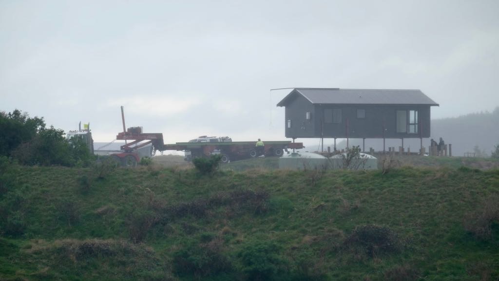 A house elevated on stilts is being transported on a large flatbed trailer across a hilly, grassy landscape.