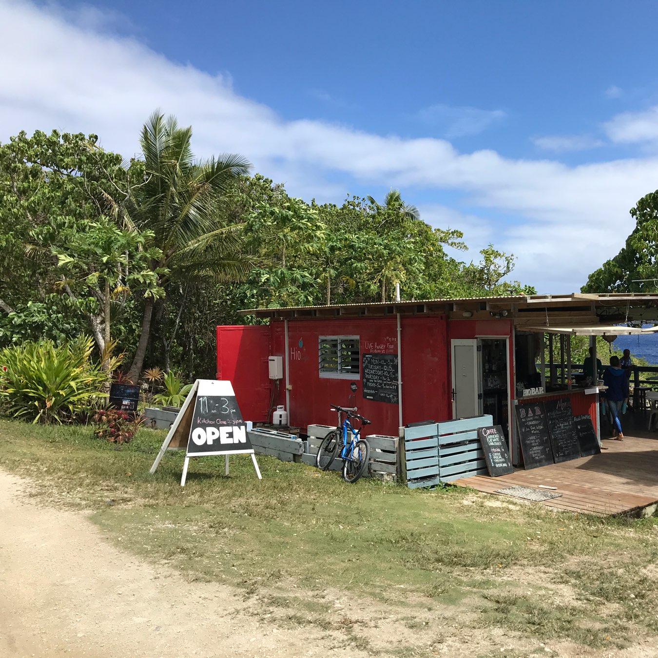 A small red roadside cafe surrounded by greenery with an OPEN sign and a blue bicycle parked nearby under a partly cloudy sky.