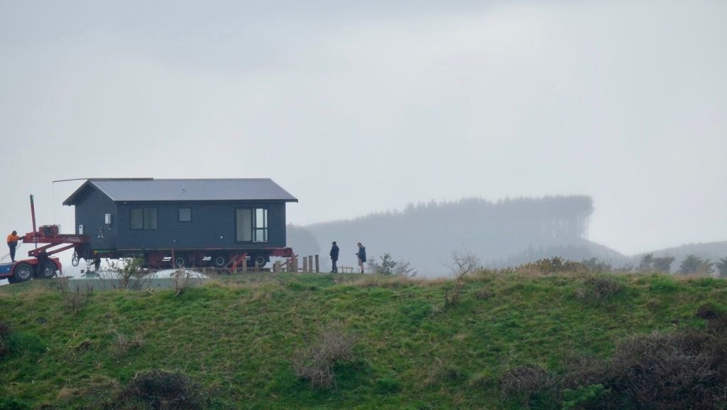 A small house is being transported on a large red vehicle along a grassy hill under a cloudy sky.