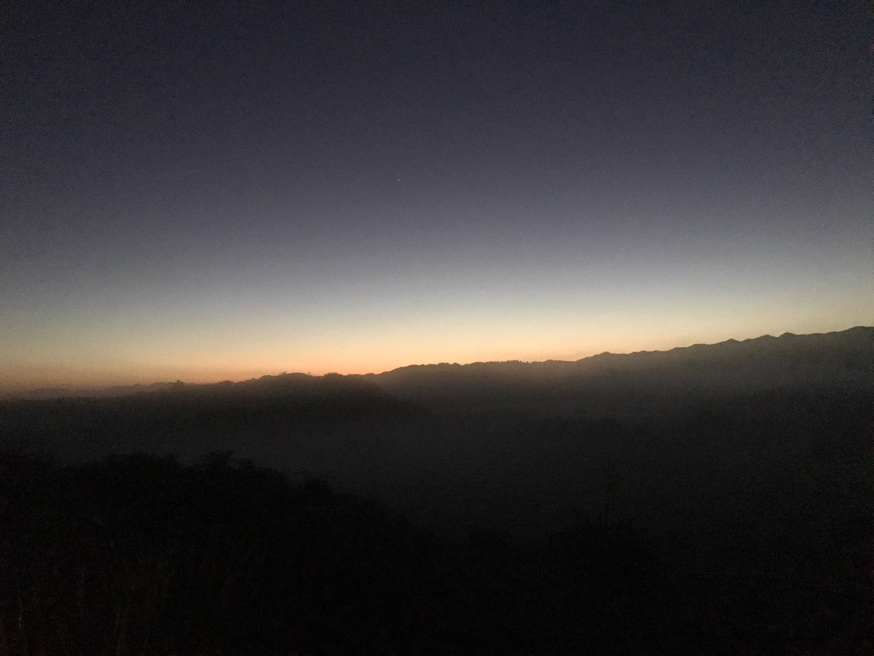 A serene mountain landscape is seen at dusk with the horizon glowing softly as the sun sets behind the peaks.
