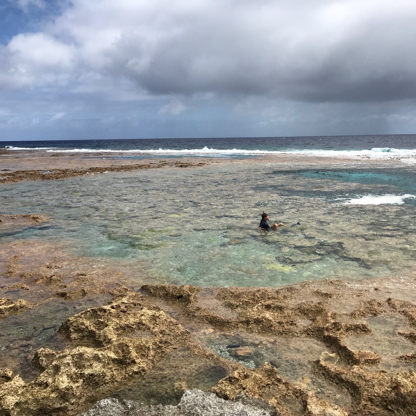 A person wearing snorkeling gear is swimming in shallow, clear waters near a rocky coastline under a cloudy sky.