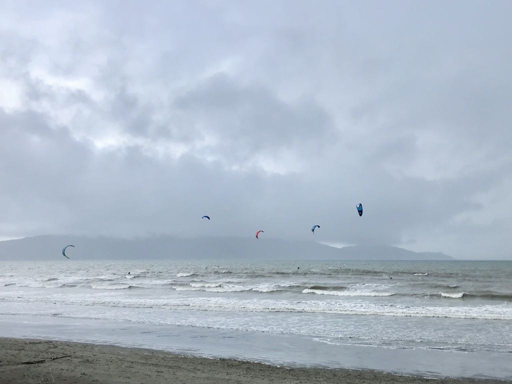 Several kite surfers are riding the waves near a cloudy and misty shoreline with distant hills visible on the horizon.