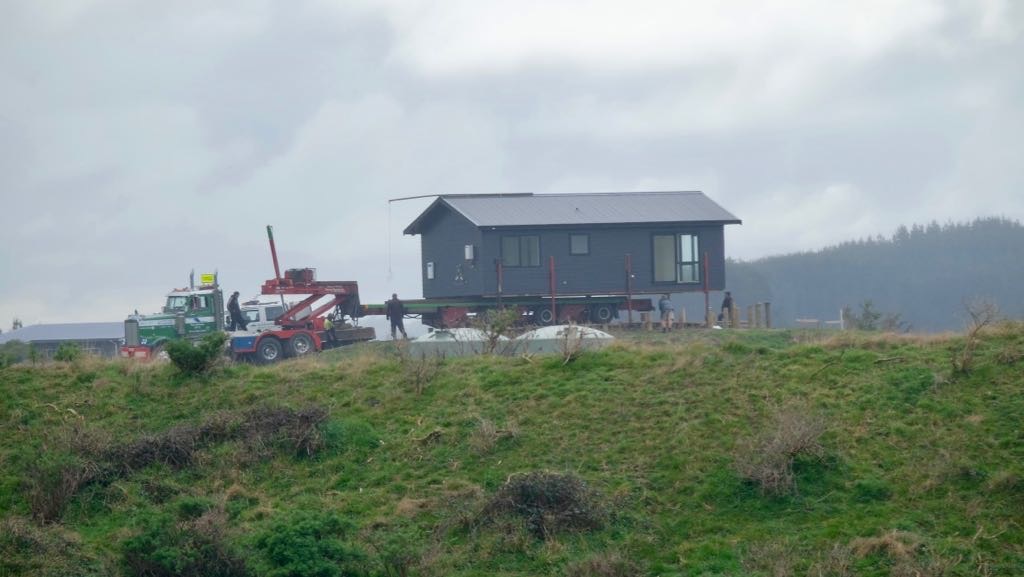 A small house is being transported on a flatbed trailer with the help of trucks and workers on a cloudy day.