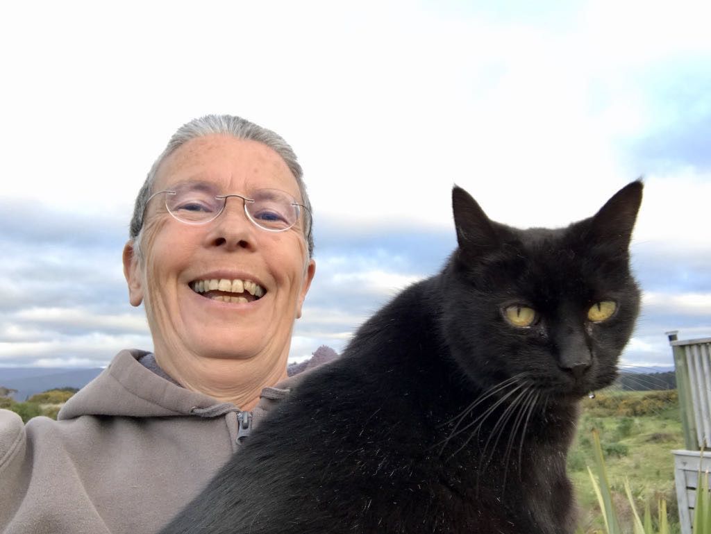 A smiling person wearing glasses and a gray hoodie is holding a black cat outdoors with a cloudy sky and some greenery in the background.