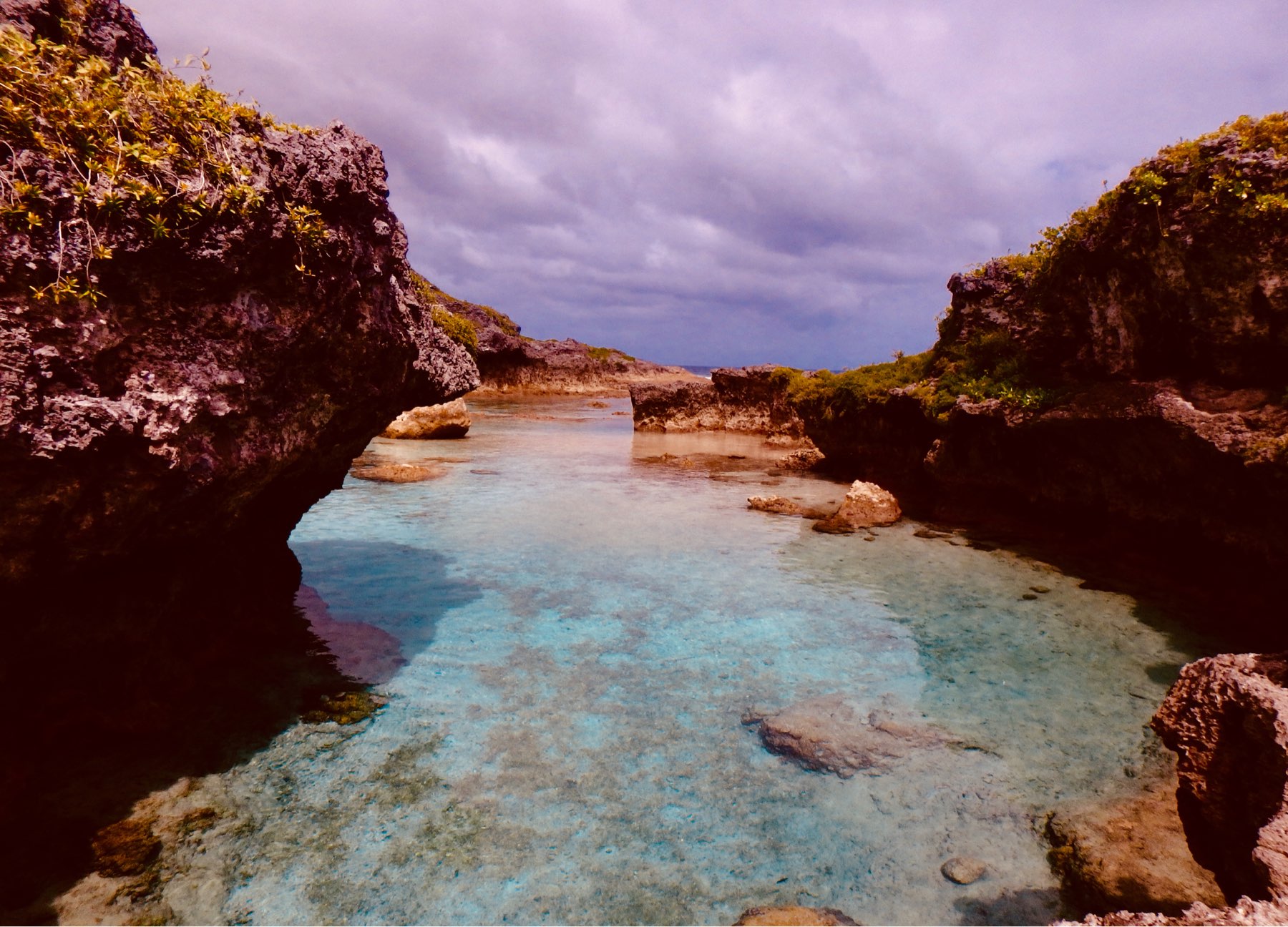 A clear, shallow body of water is surrounded by rocky cliffs under a cloudy sky.