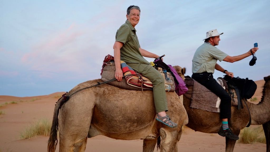 Two people are riding camels in a desert landscape during what appears to be early evening or late afternoon.