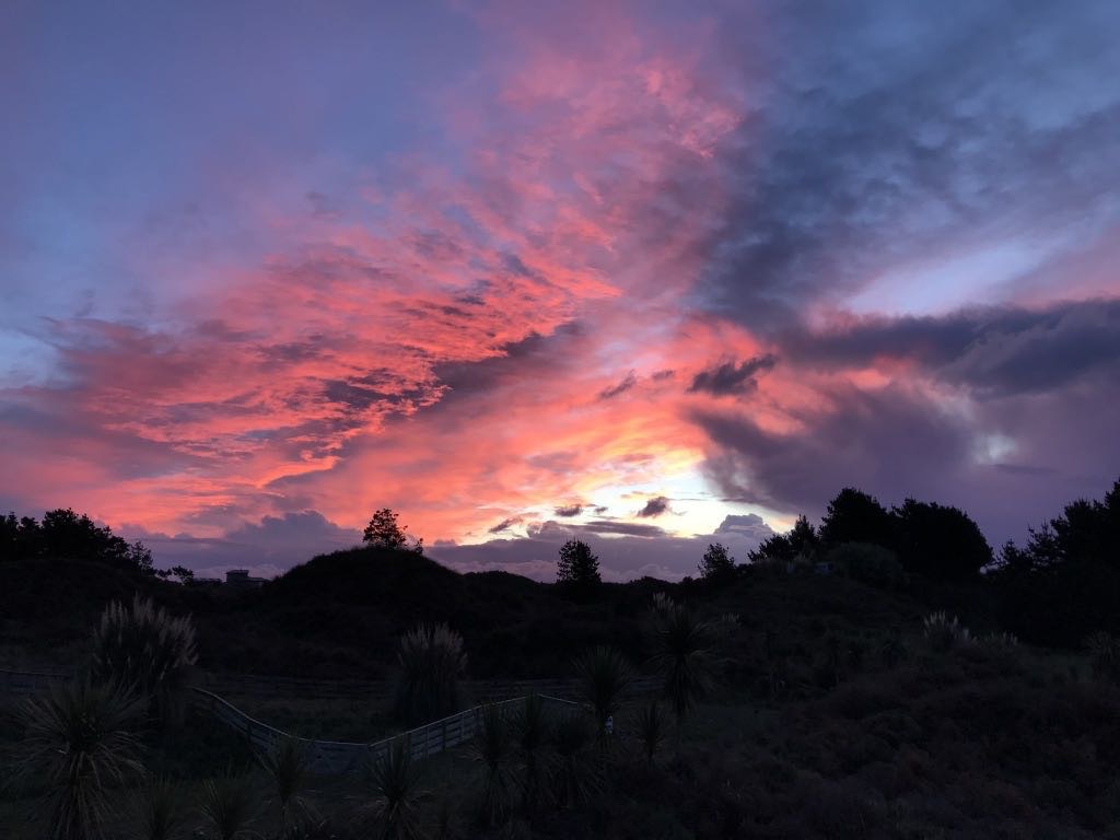 A vibrant sunset with pink and purple clouds lights up the sky above a silhouetted landscape with trees and hills.