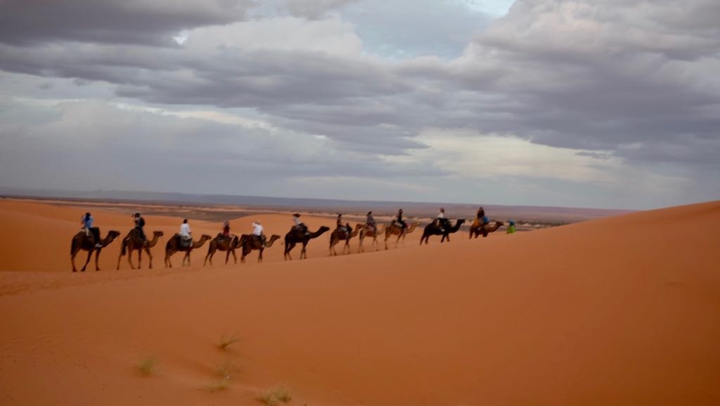 A group of people riding camels is traveling across a vast desert landscape under a cloudy sky.
