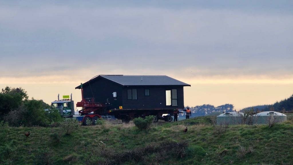 A black house is being transported on a large vehicle across a grassy field under a cloudy sky.