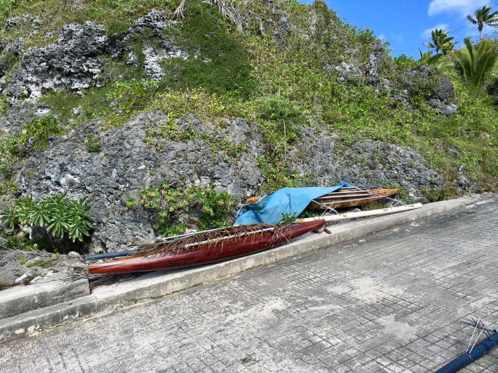 A traditional wooden boat covered partially with a blue tarp is resting on a sloped concrete surface near a rocky, grass-covered hillside.