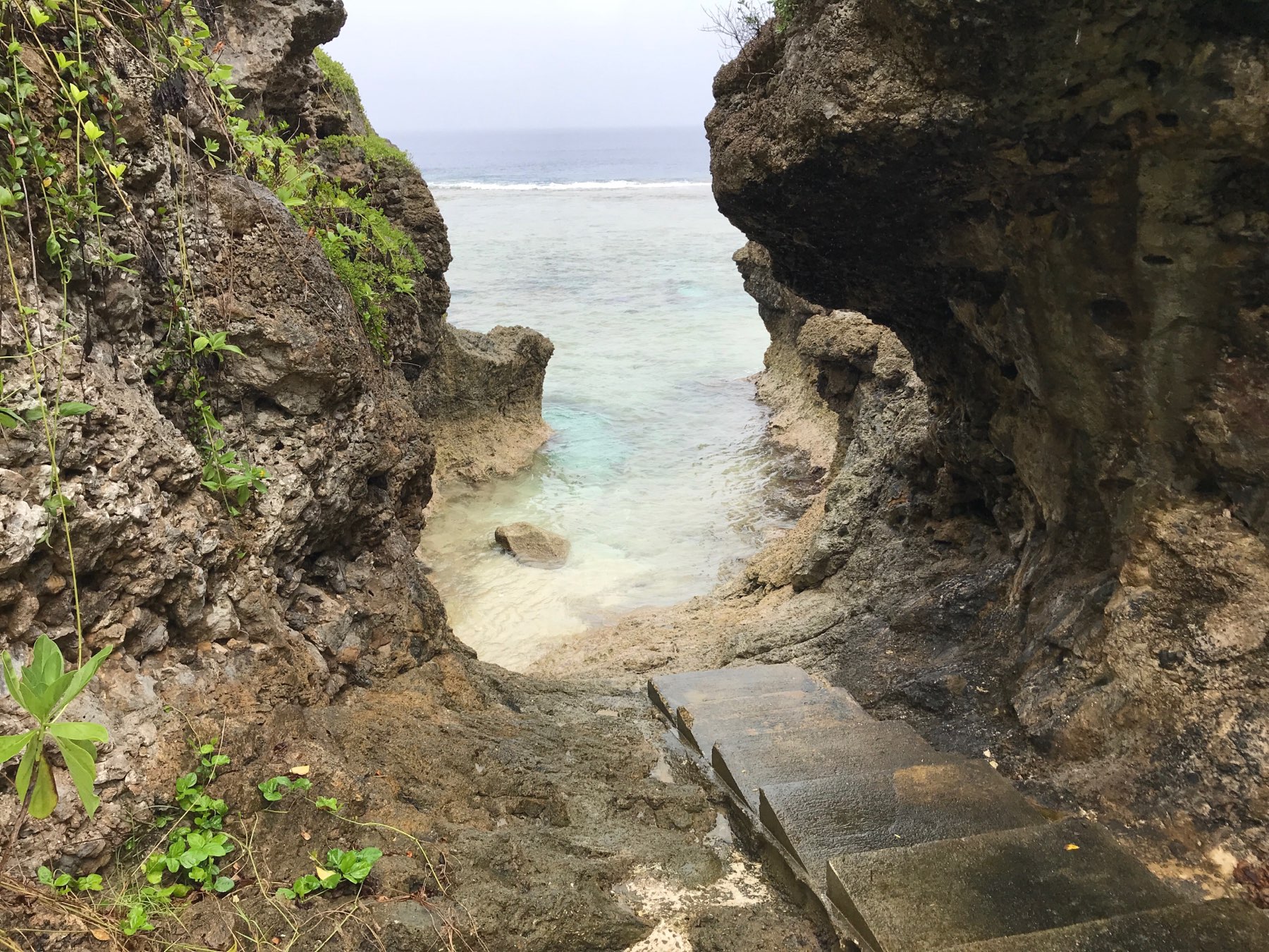 A narrow rocky pathway with small steps leads down to a clear, shallow coastal area surrounded by rugged cliffs.