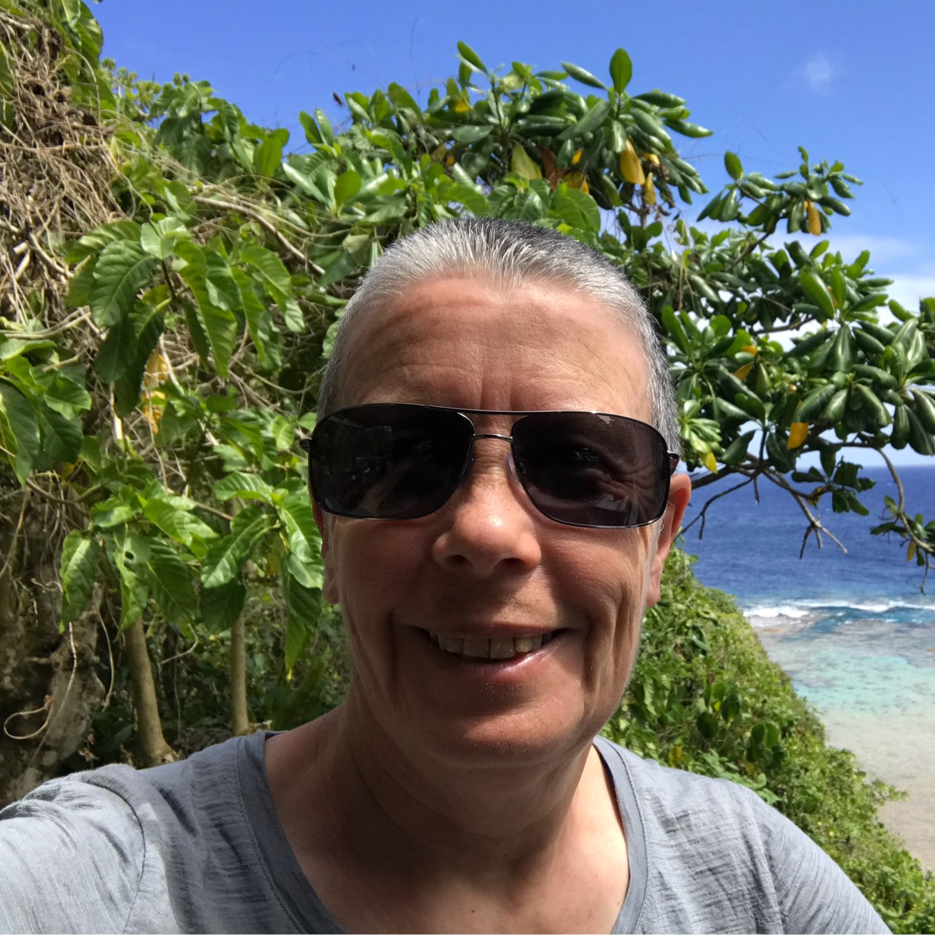 A person wearing sunglasses and a gray shirt is smiling with lush green foliage and a blue ocean visible in the background.