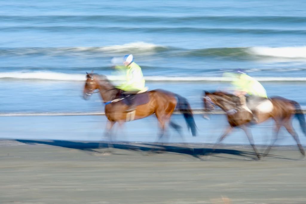 Two dark brown horses, with riders, on the beach. The horses legs are blurry.