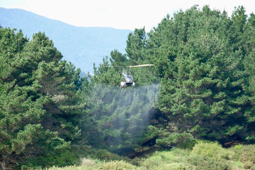 Another angle: Low-flying helicopter with trees in the backgtound and spray coming from booms below. 