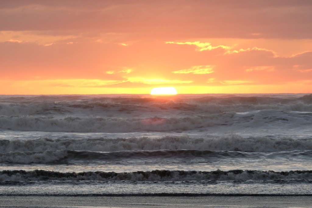 Orange sky with sun sinking below the horizon and waves like shelving in the foreground. 