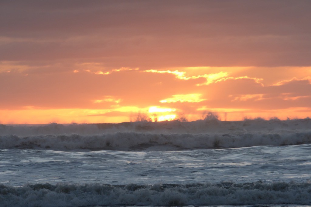 Sea in foreground, dramatic orange sky, sea spray rising high on the horizon. 