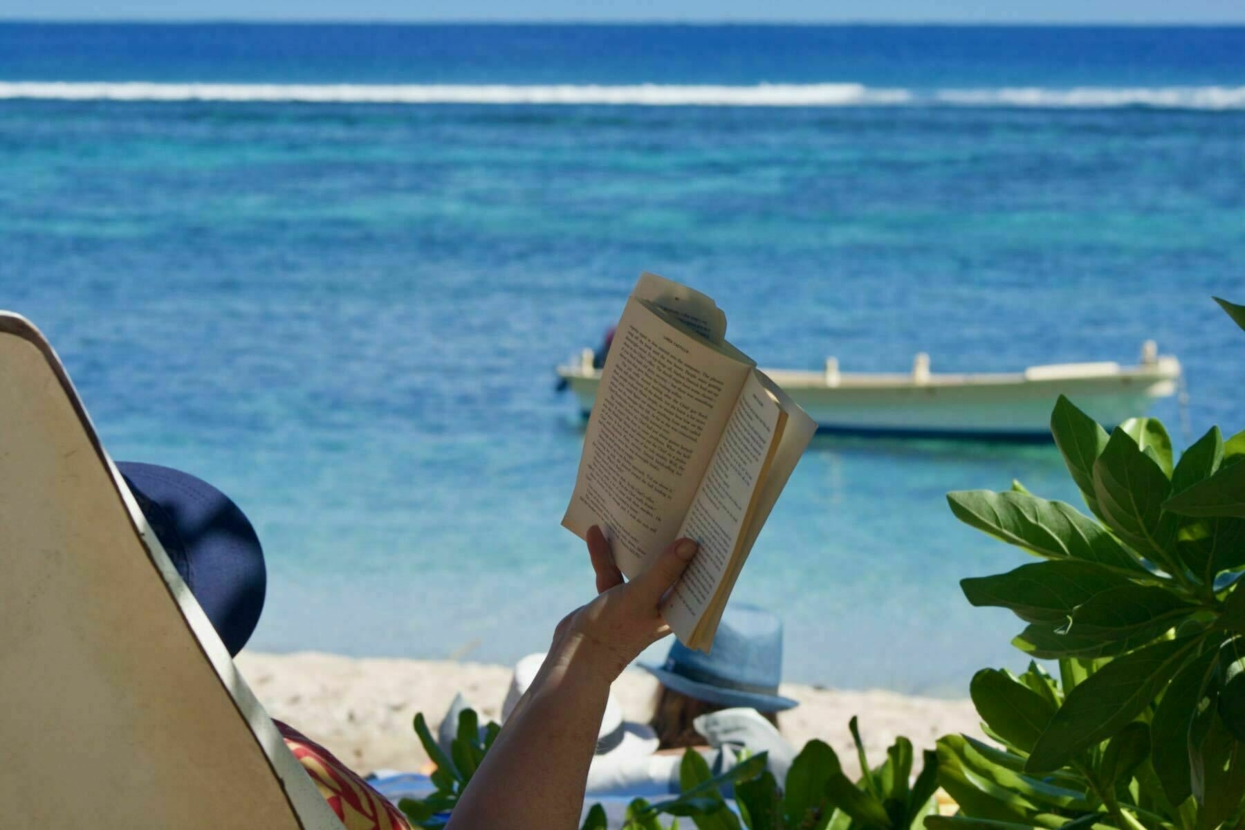 Rear view of an obscured person in a deckchair on the beach, arm extended with book, blue sea behind. 
