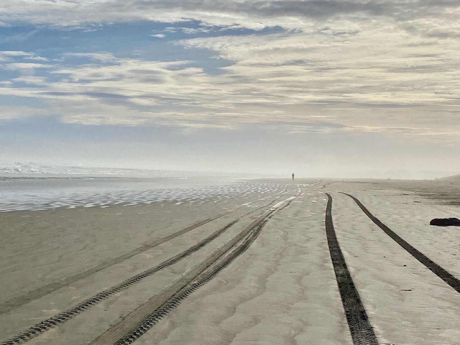 Tire tracks along the beach leading to the distance with a runner receding into the mist. 