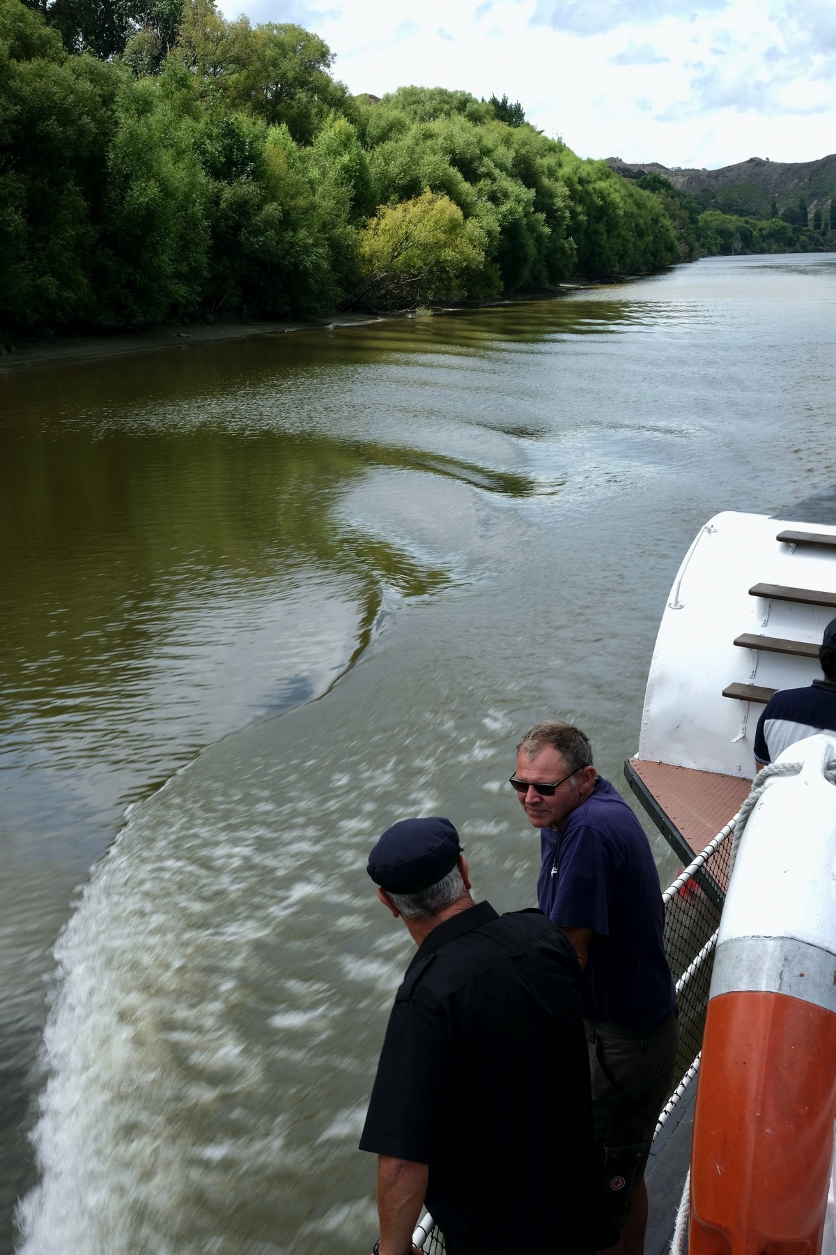 Two men at the side of the boat look at the wake. 