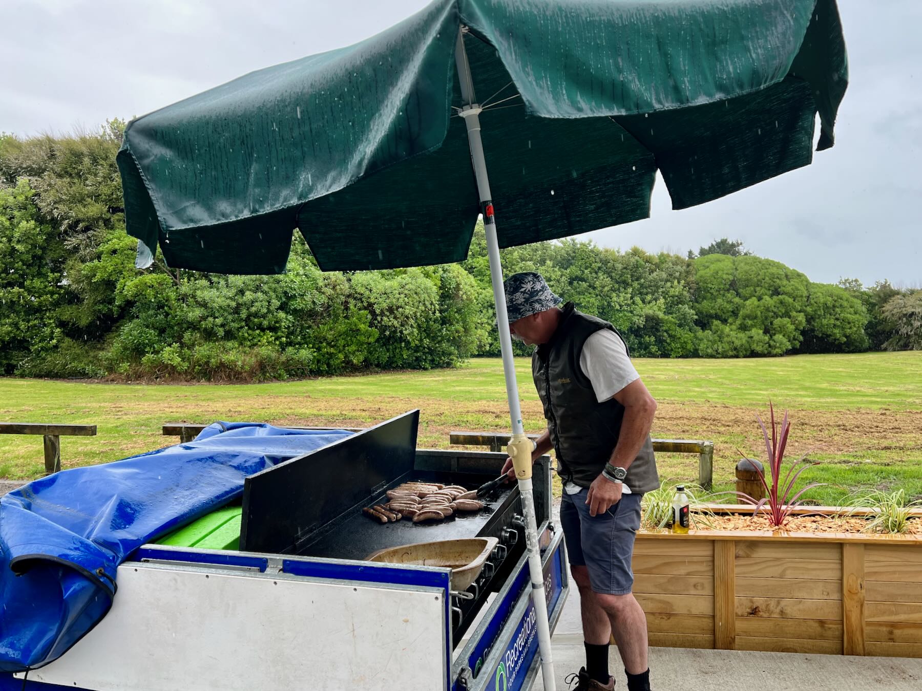 Man stands under a big umbrella cooking sausages on a BBQ in a trailer. 