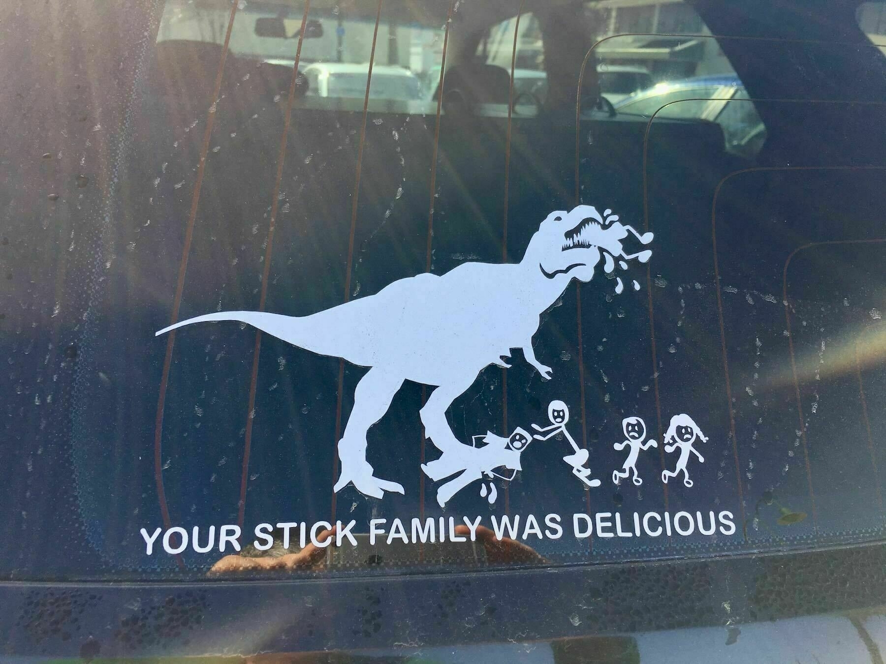 Car sticker that shows a dinosaur eating stick figures, captioned Your stick family was delicious.