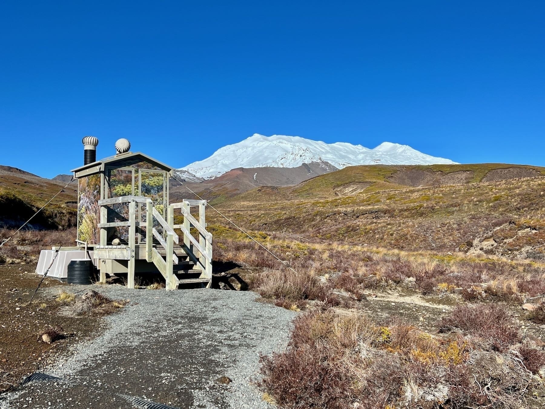 Toilets at the top of Taranaki Falls track, with the snowy top of Mt Ruapehu behind and open scrub land around. 