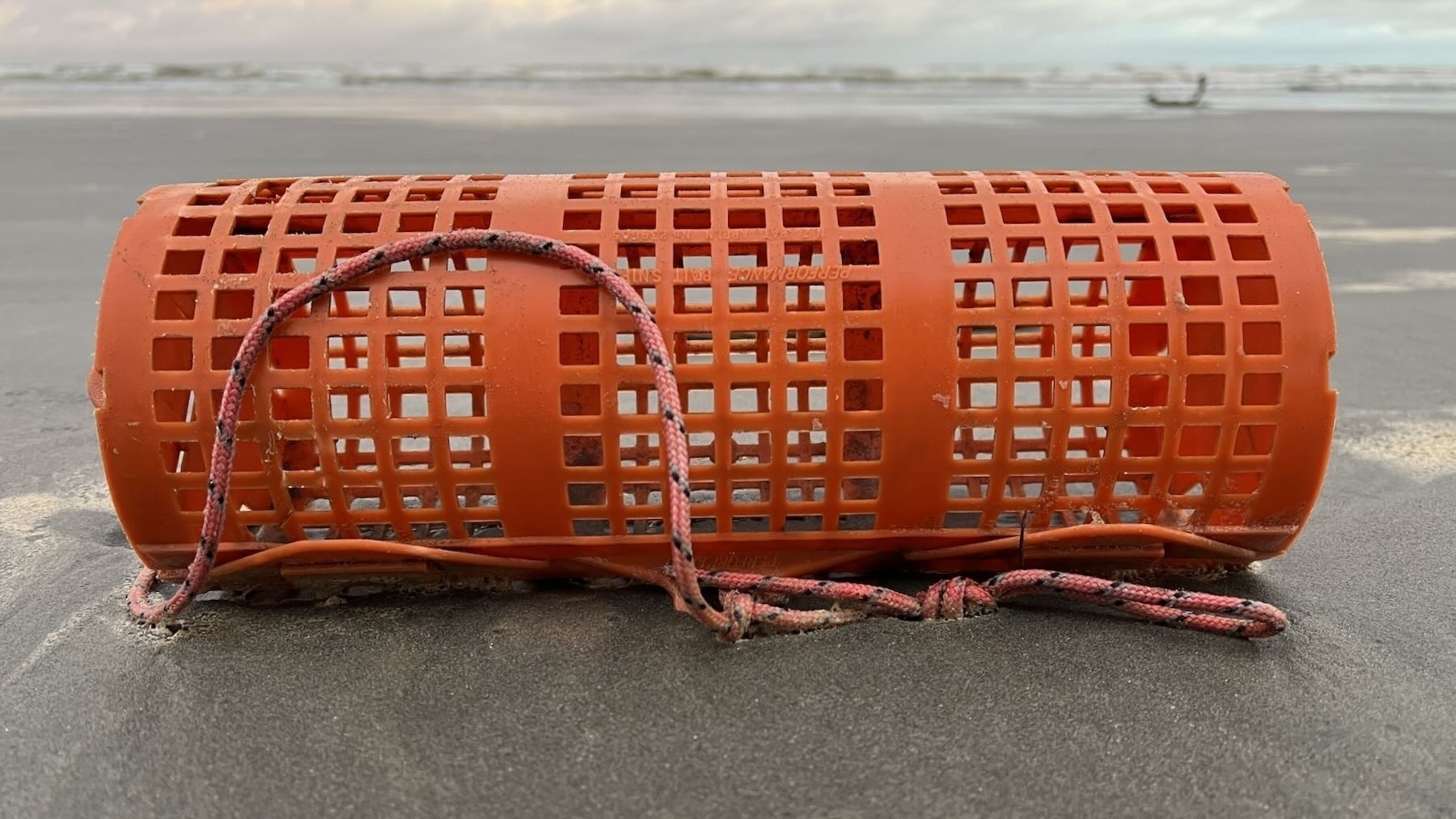 Orange cyclinder made of plastic, with small holes to allow bait to escape and fish to feed. 