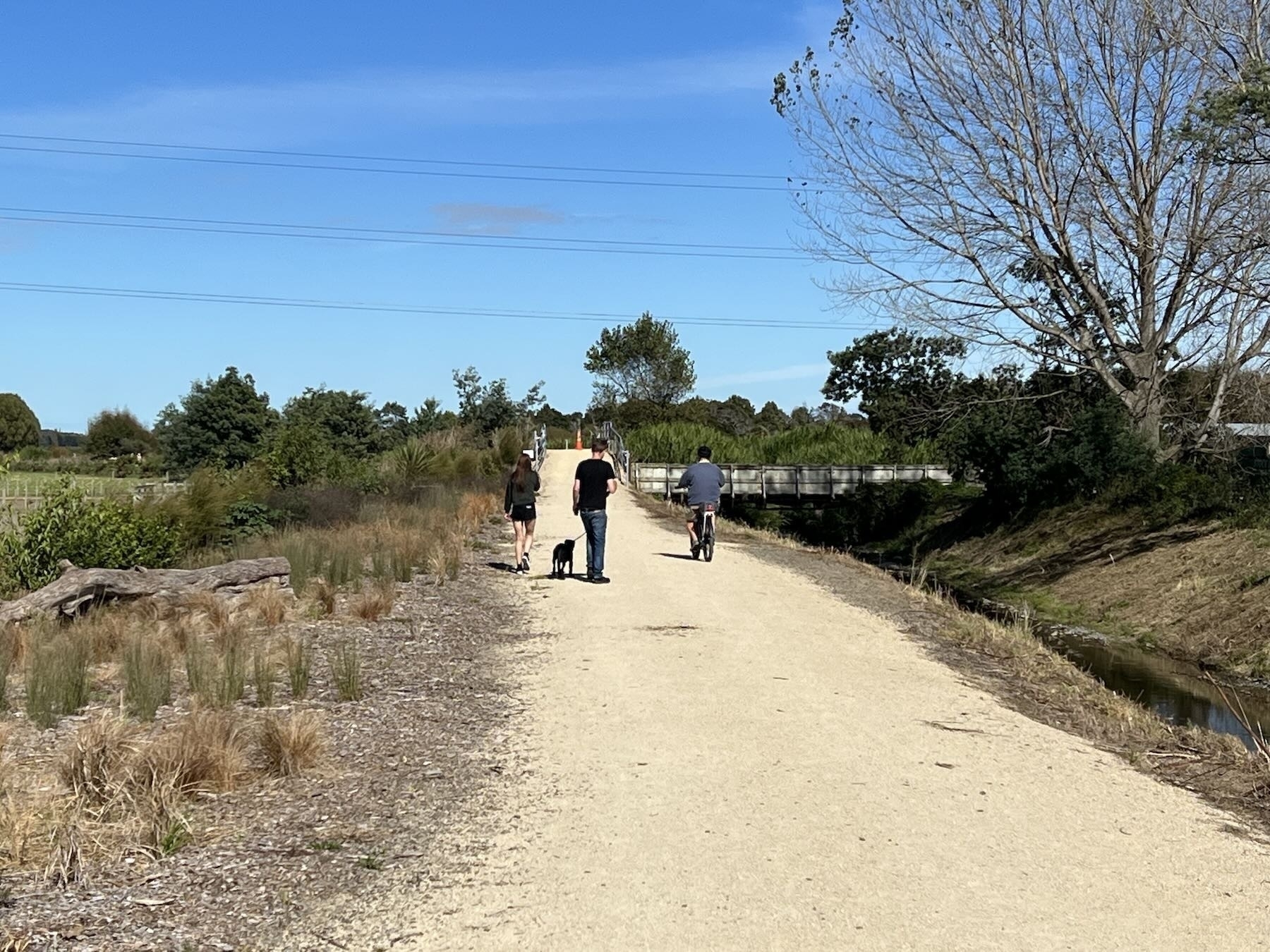 A flat formed dirt path with two people walking a dog and a cyclist. 