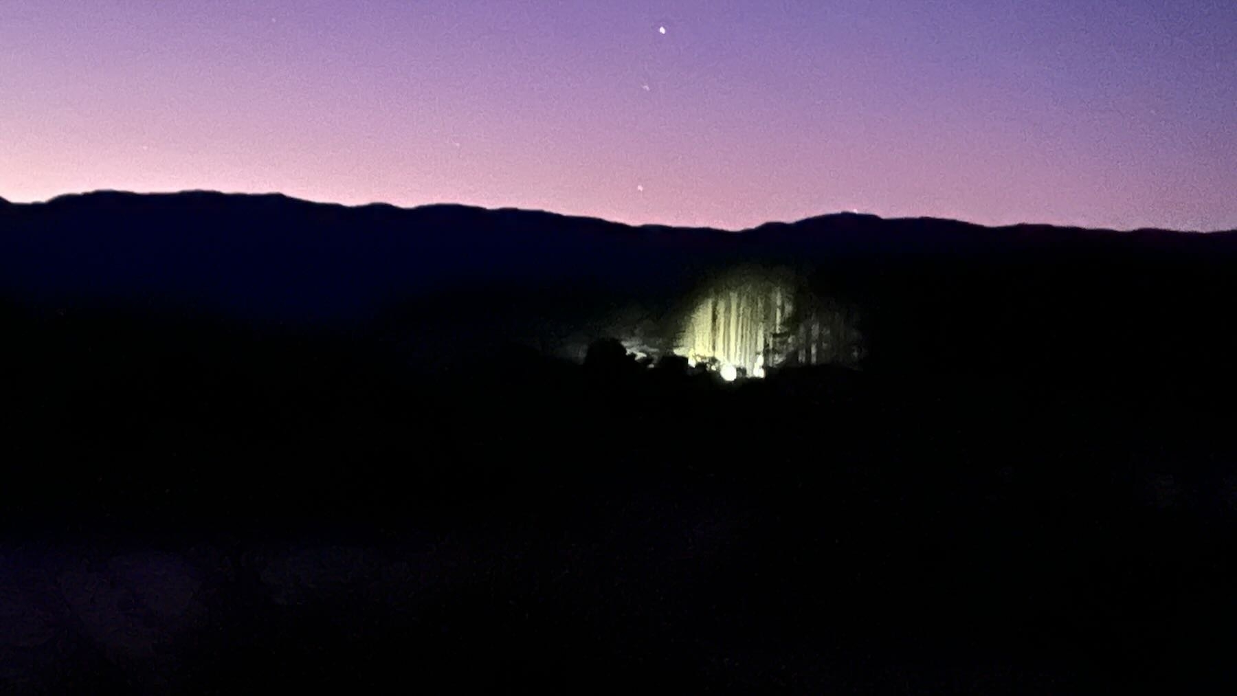 A dawn pink sky, with a dark mass below in while a puddle of light is showing pine trees. 