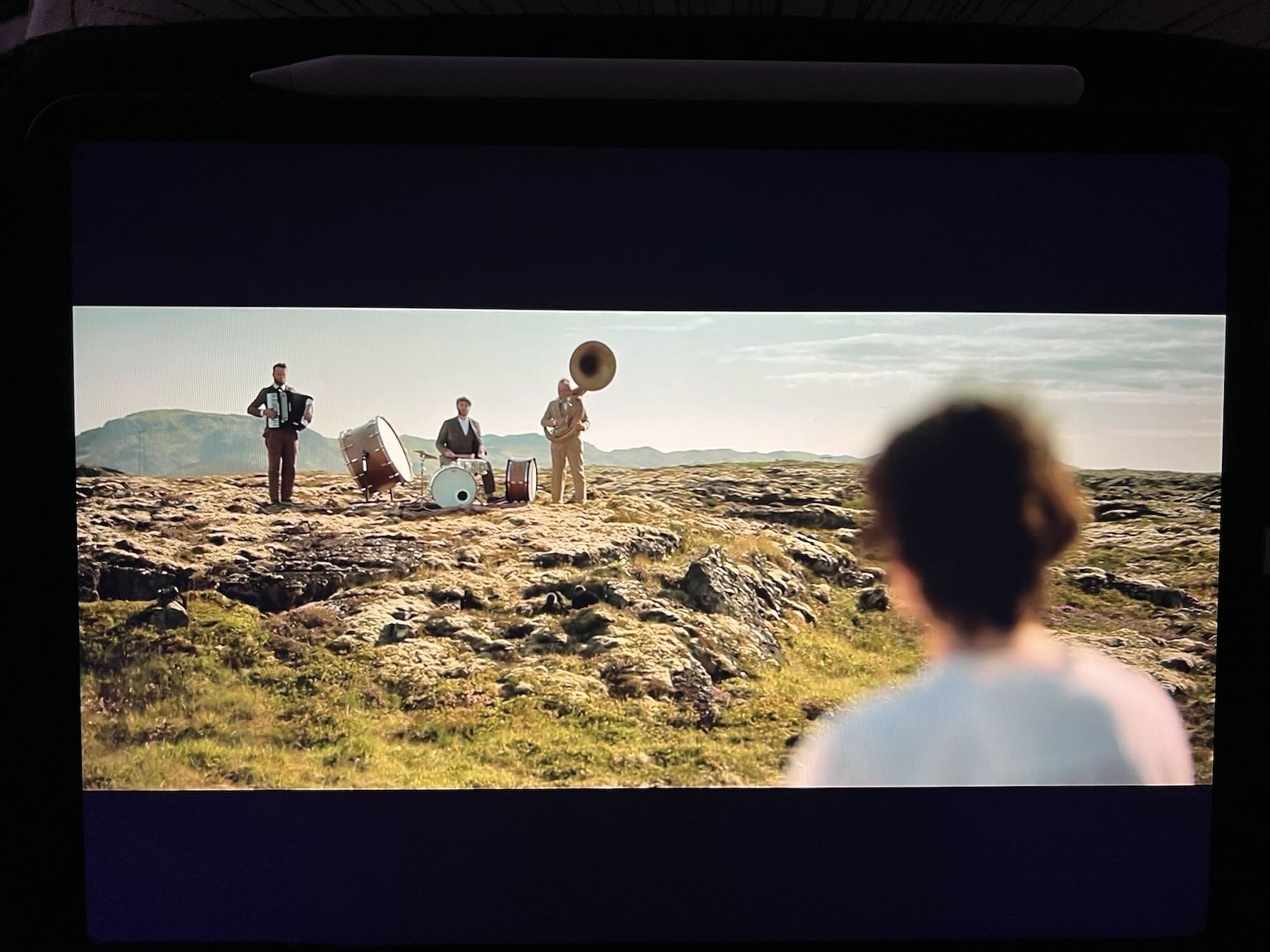 Three member band plays in Icelandic landscape. Woman in foreground is watching.