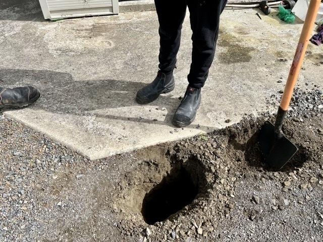 A pair of booted legs beside two holes in a gravel driveway. 